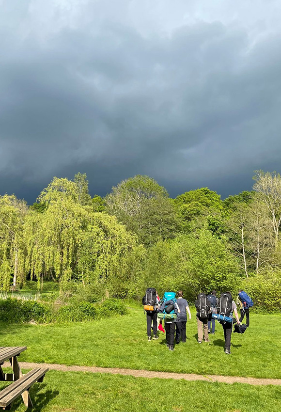 group of hikers with backpacks walking through lush green field under a dark cloudy sky enjoying the 11 nature trails