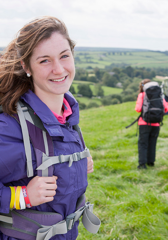 smiling young woman hiking on a green hillside wearing a purple jacket with a backpack two hiking companions in the background exploring nature during an outdoor adventure 5 amazing trails