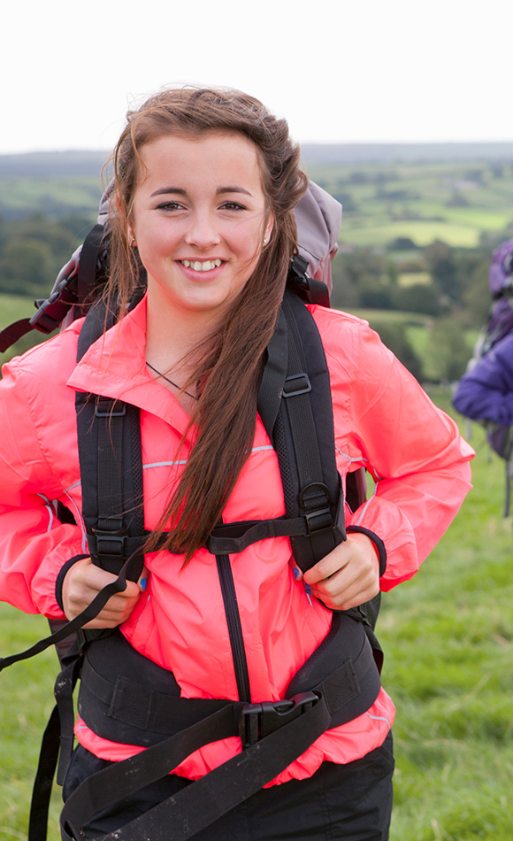 young woman smiling while wearing a bright pink jacket and a backpack in a green landscape suitable for outdoor adventures and hiking 4 essential gear items