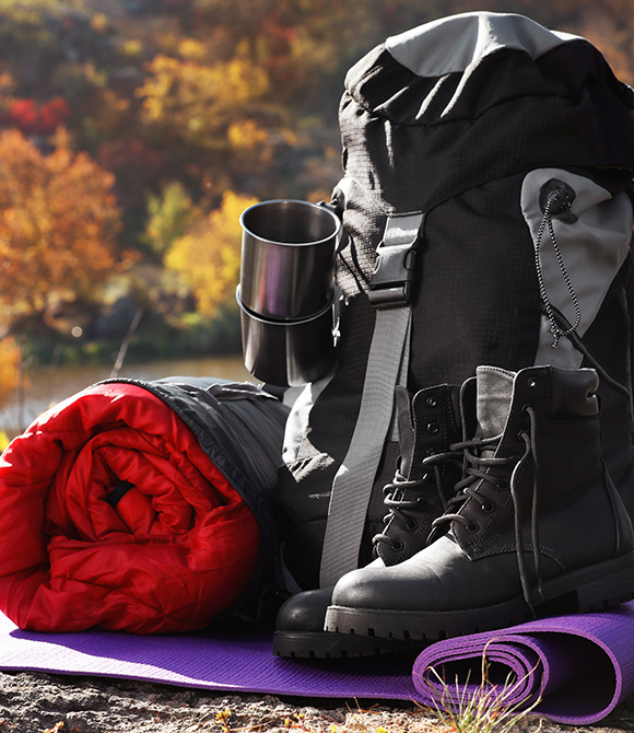 Camping gear including a black backpack with a silver mug sleeping bag and sturdy black boots on a purple mat surrounded by autumn foliage ideal for outdoor adventures and comfort.